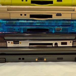 May include: A stack of Nintendo DS handheld gaming consoles in various colors, including lime green, gold, transparent blue, black, and dark green. The consoles are stacked on top of each other, showing the top and side profiles.