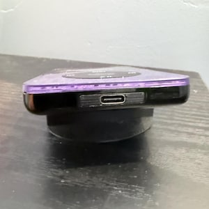 May include: A purple digital music player featuring a black circular navigation wheel on its top surface and a centered USB-C charging port located on the front edge of the device, which is supported by a black circular stand.