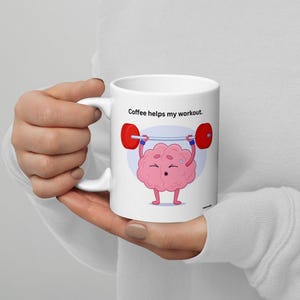 Op de afbeelding: Een witte koffiemok met een cartoonbrein dat een halterstang optilt. De tekst op de mok luidt "Coffee helps my workout."