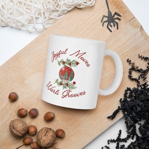 May include: White ceramic mug with a red and green Christmas-themed design. The mug features the text "Joyful Moves Warli Grooves" and a stylized illustration of a Christmas ornament with a winter scene.
