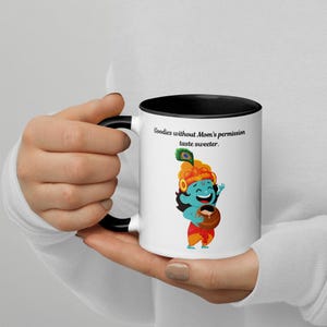 Può includere: Una tazza da caffè in ceramica bianca con bordo e manico neri. La tazza presenta un'illustrazione a fumetti di una divinità indù dalla pelle blu che tiene un vaso di latte. Il testo "Goodies without Mom's permission taste sweeter." è stampato sulla tazza.