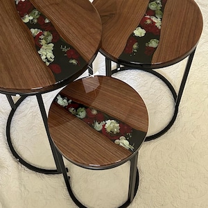 May include: A set of three round nesting tables featuring dark brown wood tops with a central black resin inlay. The resin sections are embedded with preserved red and white flowers. Each table is supported by a black metal frame base.