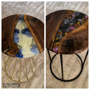May include: Two round wooden side tables with resin tops featuring pressed flowers. The table on the left has a white resin top with blue flowers and a gold metal base. The table on the right has a black resin top with purple, pink, and yellow flowers and a black metal base.