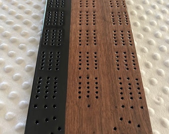 May include: A wooden cribbage board with a black resin inlay on one side. The board is rectangular with rounded edges and features numerous small holes for peg placement. The other side is a natural wood color.
