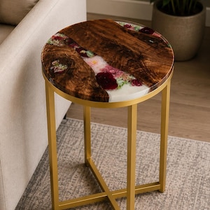 May include: A round side table featuring a natural wood slab top with a dark brown grain, embedded with translucent white resin and preserved colorful flowers. The table is supported by a gold-colored metal frame with four straight legs.