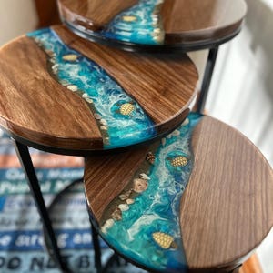 Puede incluir: Juego de tres mesas auxiliares redondas. Las tapas de las mesas son de madera con un río de resina azul, conchas y detalles dorados. Las mesas tienen patas de metal negro. El texto de fondo dice "DO NOT BE DISCOURAGED".