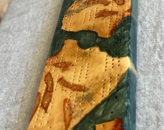 May include: A wooden cribbage board with a unique design. The board features a light brown surface with dark green and red accents, and a blue resin border. It has numerous holes for pegs and includes four metal pegs.