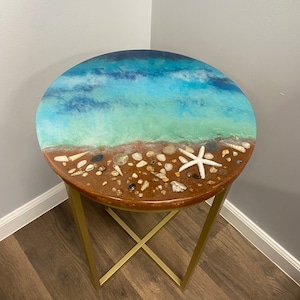 Puede incluir: Mesa auxiliar redonda con base de metal dorado y tapa de resina con una escena de océano azul y verde con conchas y una estrella de mar.