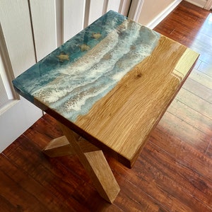May include: A wooden end table with a resin top featuring a blue and white ocean wave design with three gold sea turtles. The table has a simple X-shaped base.