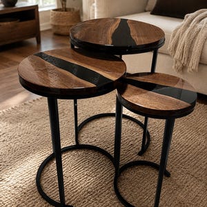 May include: A set of three round nesting tables featuring natural wood grain tops with a glossy black resin inlay running through the center. Each table is supported by a black metal frame with a curved base, creating a tiered arrangement.