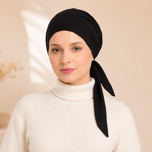 May include: A solid black head wrap made of smooth fabric that covers the hair and ties at the side with two long, pointed ends. The head wrap is worn over a cream-coloured, ribbed knit jumper featuring a high, folded turtleneck collar.