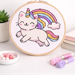 May include: A cross-stitch embroidery hoop featuring a whimsical design of a cat with a unicorn horn and a rainbow. The cat is light pink, and the rainbow has multiple colors. The hoop is made of wood and is displayed on a white surface.