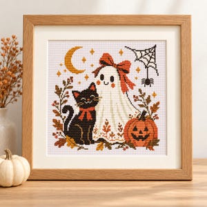 May include: A framed cross-stitch embroidery featuring a black cat with a red bow, a white ghost wearing a red bow, and an orange jack-o'-lantern. The design includes a yellow crescent moon, a black spider web, and autumn leaves in a light wood frame.