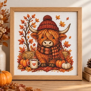 Könnte beinhalten: Eine Kreuzstichstickerei in einem hellen Holzrahmen zeigt ein flauschiges, rotbraunes Hochlandrind mit einer roten Strickmütze und einem karierten Schal, umgeben von herbstlichen Blättern, Kürbissen und einer weißen Tasse mit Herzmotiv.