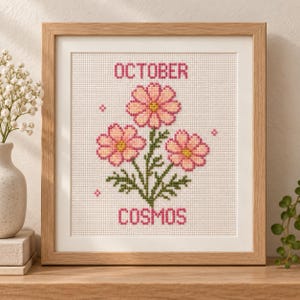 Può includere: Un ricamo a punto croce quadrato in una cornice di legno chiaro raffigurante tre fiori di cosmo rosa con centri gialli, steli e foglie verdi. Il testo OCTOBER è ricamato in rosa sopra i fiori e COSMOS è ricamato in rosa sotto di essi.