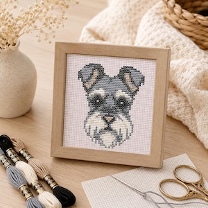 Könnte beinhalten: Ein quadratischer Holzrahmen mit einer Kreuzstichstickerei eines grau-weißen Schnauzer-Hundekopfes. Das Design zeigt detaillierte Stiche in Grau-, Schwarz- und Cremetönen auf einem weißen Stoffhintergrund innerhalb des hellen Holzrahmens.