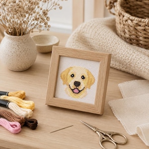 May include: A square light wood frame containing a cross-stitch embroidery of a golden retriever dog face. The dog has yellow fur, black eyes, a black nose, and a pink tongue. The embroidery is on white fabric, displayed in a simple wooden frame.