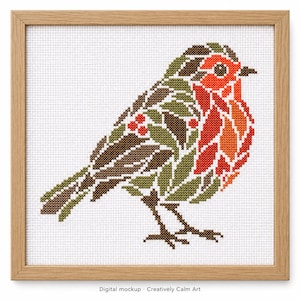 May include: A cross-stitch pattern of a robin bird in a light wood frame. The bird is composed of green leaves, brown feathers, and bright red berries on its chest and wing. The design is stitched onto white Aida fabric with a grid texture.