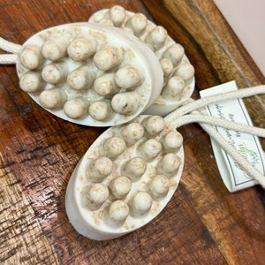 Może przedstawiać: Trzy białe owalne mydła z fakturowaną powierzchnią i przymocowaną białą sznurkiem. Mydła leżą na drewnianej powierzchni.