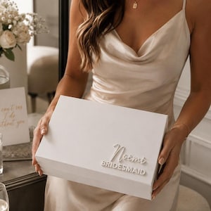 Puede incluir: Una caja de regalo rectangular blanca con el nombre Noemi en letra cursiva en relieve sobre la palabra BRIDESMAID en letras de molde. La caja es sostenida frente a una persona que viste un vestido lencero de satén color champán con cuello drapeado.
