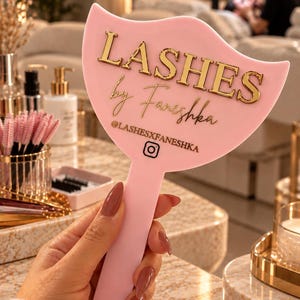 May include: A light pink handheld mirror with a curved top edge. The mirror features gold 3D lettering that reads LASHES by Faneshka, followed by the text @LASHESXFANESHKA and an Instagram icon printed in a dark color on the smooth surface.