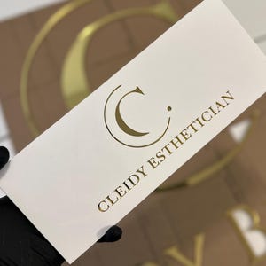 Könnte beinhalten: Eine weiße rechteckige Karte mit goldfarbenen Buchstaben und einem Logo wird von einer behandschuhten Hand gehalten. Auf der Karte steht "CLEIDY ESTHETICIAN" in Gold, mit einem goldenen "C"-Logo. Im Hintergrund ist ein größeres Schild mit goldenen Buchstaben zu sehen.