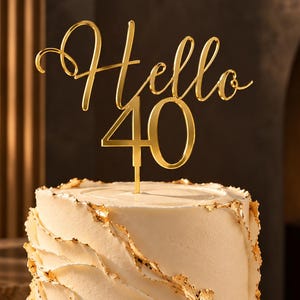 May include: A gold-colored acrylic cake topper featuring the script text Hello and the number 40, positioned atop a cylindrical white cake with textured frosting and gold leaf accents, displayed on a circular marble-patterned cake stand.