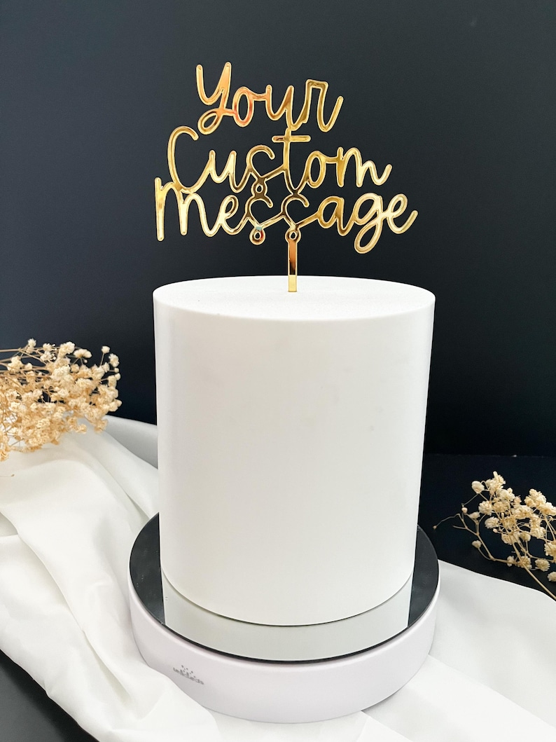 K&ouml;nnte beinhalten: Ein goldfarbener Tortenstecker aus Acryl mit dem Schriftzug Your Custom Message in Schreibschrift. Der Tortenstecker ist an einem d&uuml;nnen vertikalen Stab befestigt und steckt in einer glatten, zylindrischen wei&szlig;en Torte mit matter Oberfl&auml;che.