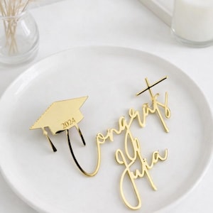 May include: Gold-colored graduation cap with the year 2024 and the words "Congrats Julia" in elegant script. The decorations are placed on a white plate, perfect for a graduation celebration.