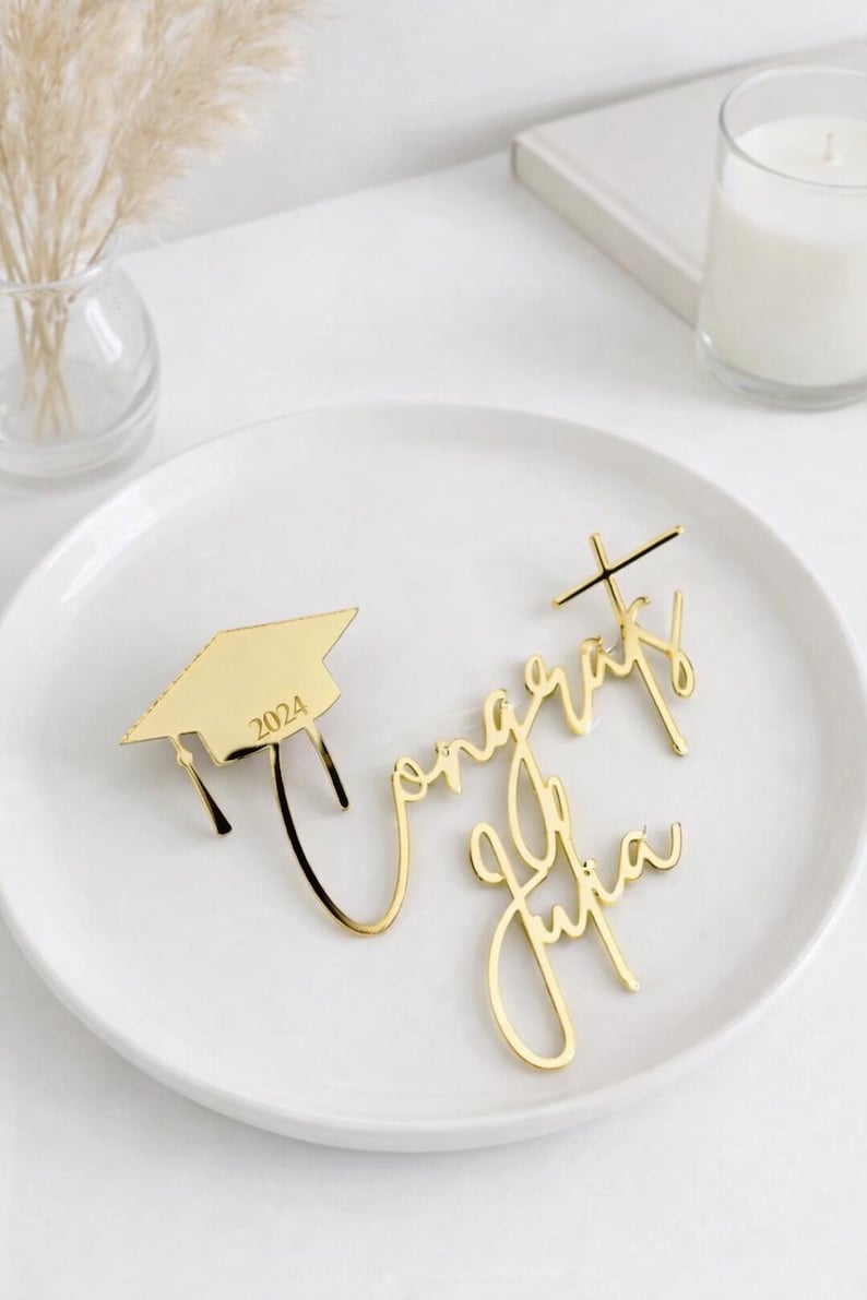 May include: Gold-colored graduation cap with the year 2024 and the words "Congrats Julia" in elegant script. The decorations are placed on a white plate, perfect for a graduation celebration.