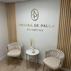 May include: A waiting area with two beige chairs with gold frames and a small round marble table with a gold frame. The wall behind the chairs has a gold logo with the letters "AP" in a circle and the text "Amanda de Paula Esthetics".