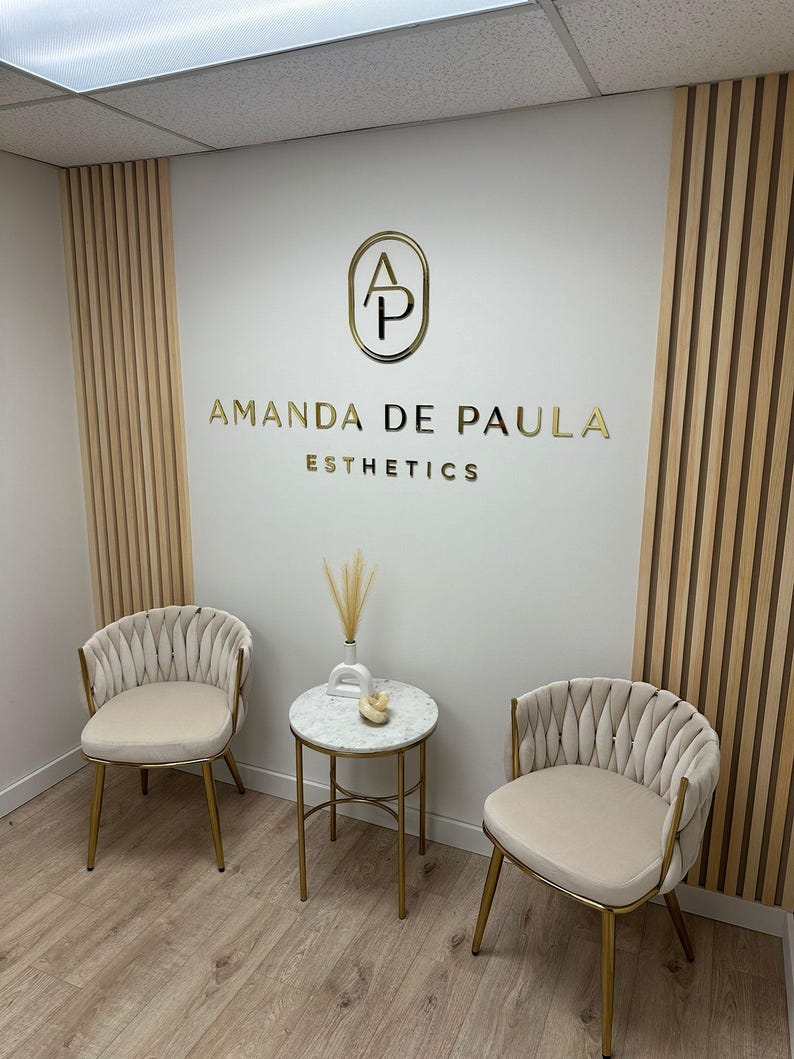 May include: A waiting area with two beige chairs with gold frames and a small round marble table with a gold frame. The wall behind the chairs has a gold logo with the letters "AP" in a circle and the text "Amanda de Paula Esthetics".