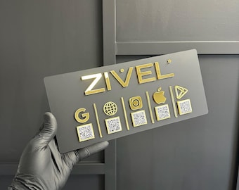 May include: A rectangular gray sign with the gold text "ZIVEL" and various gold icons, including a globe, camera, and Apple logo. The sign also features QR codes. The sign is held by a person wearing a black glove.