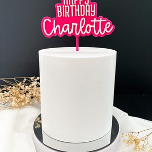 Pode incluir: Um bolo branco com um topo de bolo rosa brilhante que diz "HAPPY BIRTHAY Charlotte" em uma fonte branca em negrito. O bolo está em um pedestal branco com uma superfície espelhada, em um fundo preto. Flores secas no fundo.