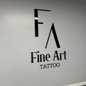 May include: A black and white sign with the text "Fine Art Tattoo" on a light gray wall.