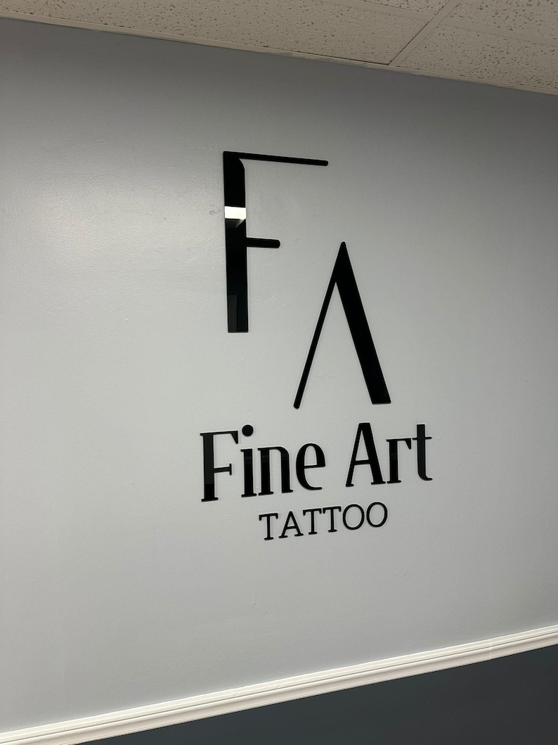May include: A black and white sign with the text "Fine Art Tattoo" on a light gray wall.