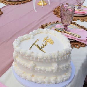 Peut inclure: Un g&acirc;teau blanc en forme de c&oelig;ur avec un gla&ccedil;age blanc et un lettrage dor&eacute; qui dit "Thirty" sur le dessus. Le g&acirc;teau est sur un support &agrave; g&acirc;teau blanc et est entour&eacute; d'une nappe rose.