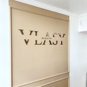 May include: A beige wall with a gold-colored sign that reads "VIACI BEAUTY". The sign is made of individual letters.