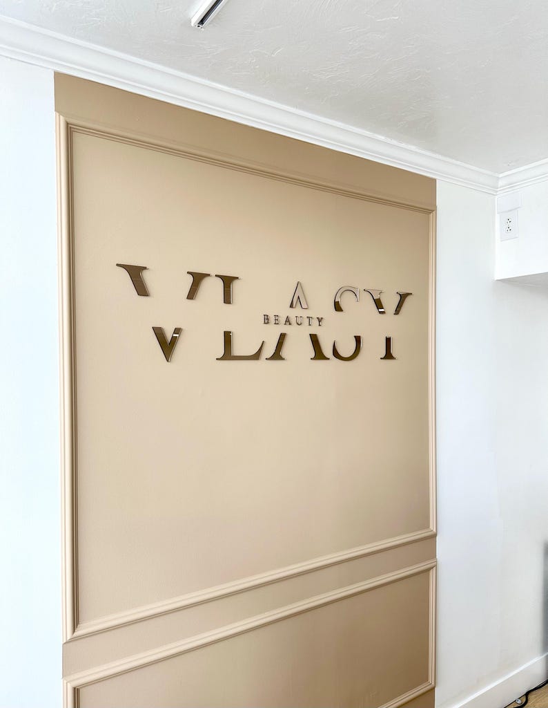 May include: A beige wall with a gold-colored sign that reads "VIACI BEAUTY". The sign is made of individual letters.