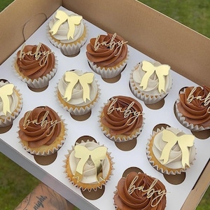 Pode incluir: Uma caixa com uma dúzia de cupcakes com cobertura de chocolate e baunilha. Alguns cupcakes têm laços dourados, outros com a palavra "baby" dourada. Os cupcakes estão em forminhas de papel brancas.