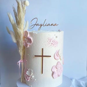 May include: White cake decorated with pink flowers and a gold cross. The cake is topped with a gold name topper that says "Jayliana".