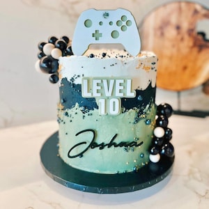 Puede incluir: Una tarta redonda con glaseado blanco, verde y negro, decorada con un mando de videojuego blanco. La tarta tiene las palabras "LEVEL 10" y el nombre "Joshua". Decoraciones de bolas blancas y negras en el lateral.