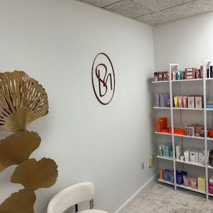 May include: A white room with a white chair and a gold decorative piece. The room has a white ceiling with square tiles. There is a shelf unit with various products on it. A brown circle with the letters "BN" inside is on the wall.