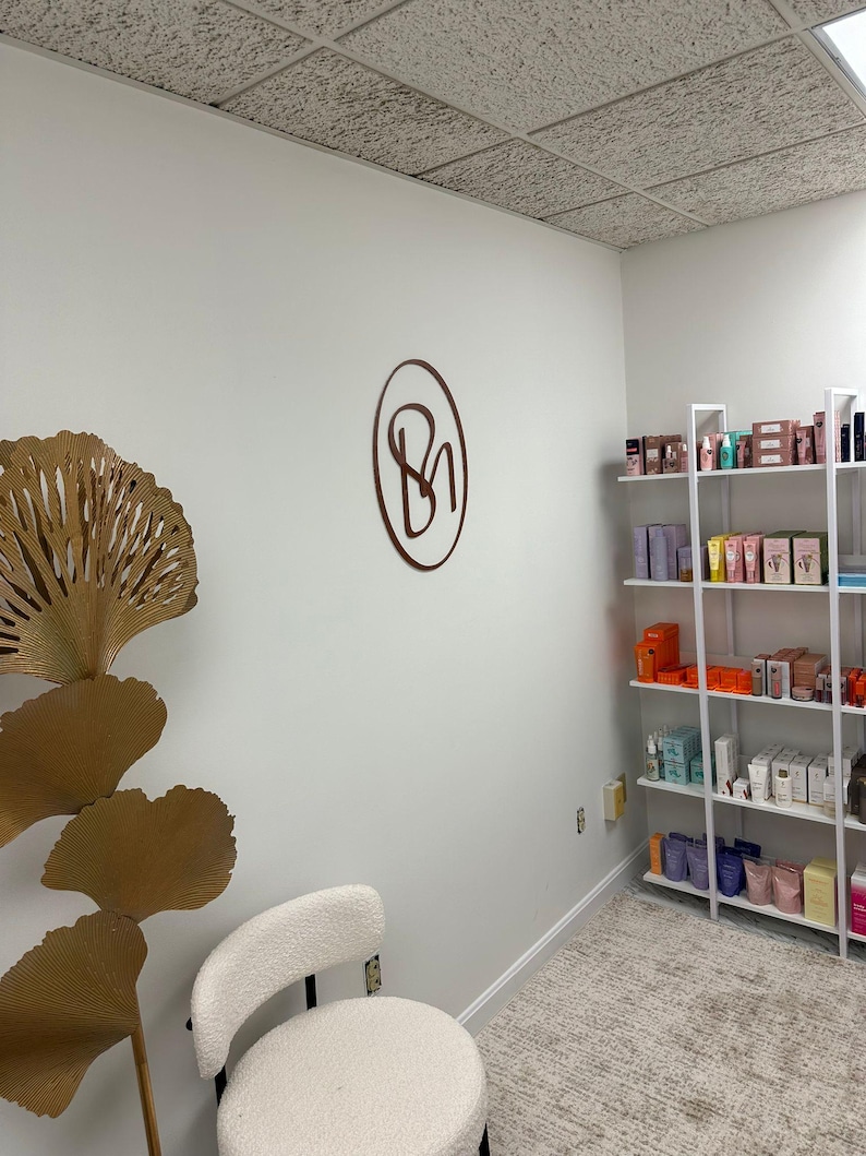 May include: A white room with a white chair and a gold decorative piece. The room has a white ceiling with square tiles. There is a shelf unit with various products on it. A brown circle with the letters "BN" inside is on the wall.