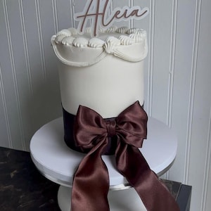 Puede incluir: Un pastel blanco con detalles marrones, decorado con un gran lazo y un adorno que dice "BABY Aleia". El pastel está sobre un soporte blanco, ideal para un baby shower u otra celebración.