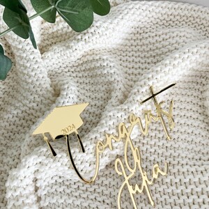 May include: Gold-colored graduation cake toppers. One is a mortarboard with the year 2024, and the other says "Congrats Julia" in cursive. The toppers are on a cream-colored knitted blanket with green leaves in the background.