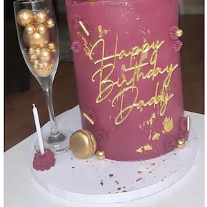 May include: A burgundy cake decorated with gold lettering that says "Happy Birthday Daddy". The cake is decorated with gold sprinkles, gold macarons, and gold beads. There is a champagne flute filled with gold beads and a white candle on a cake stand.