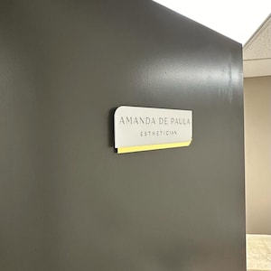 Pu&ograve; includere: Un cartello bianco con angoli arrotondati su una parete grigia. Il cartello recita "Amanda de Paula Estetista".