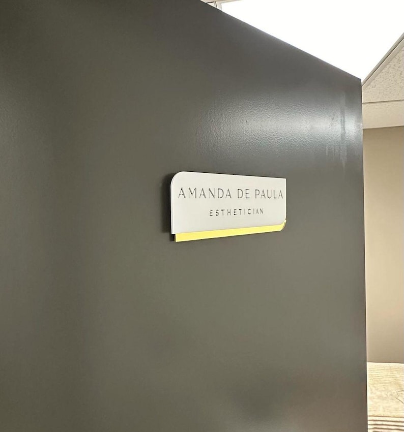 Pu&ograve; includere: Un cartello bianco con angoli arrotondati su una parete grigia. Il cartello recita "Amanda de Paula Estetista".