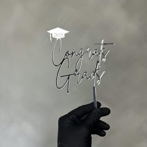 May include: A silver-colored cake topper with the words "Congrats Grads" in a script font. The year "2025" is above the words, inside a graduation cap. The topper is held by a gloved hand.